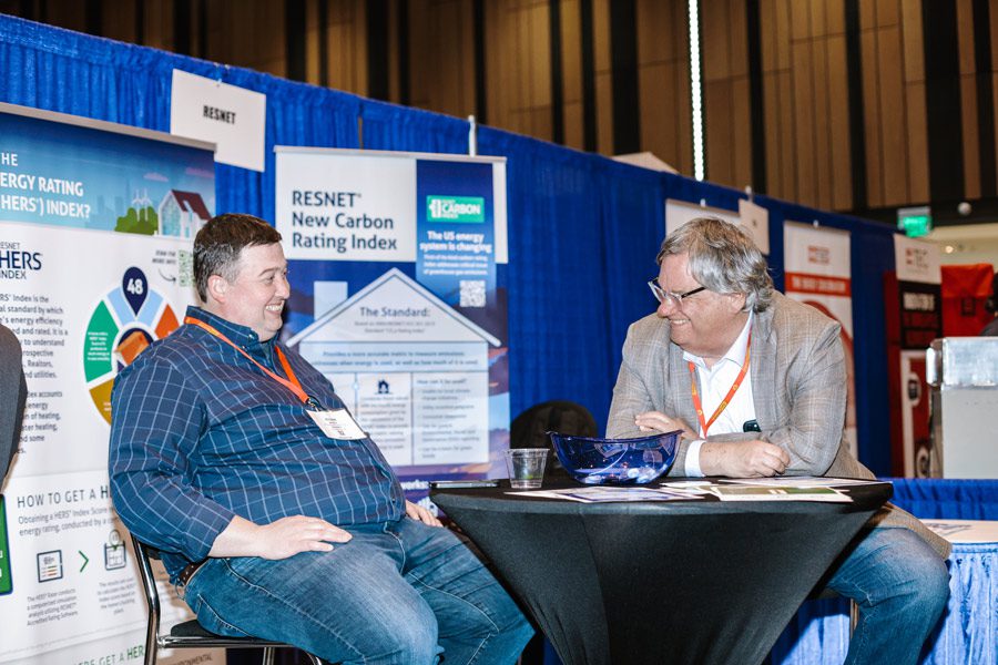 Two attendees sit in front of a booth smiling