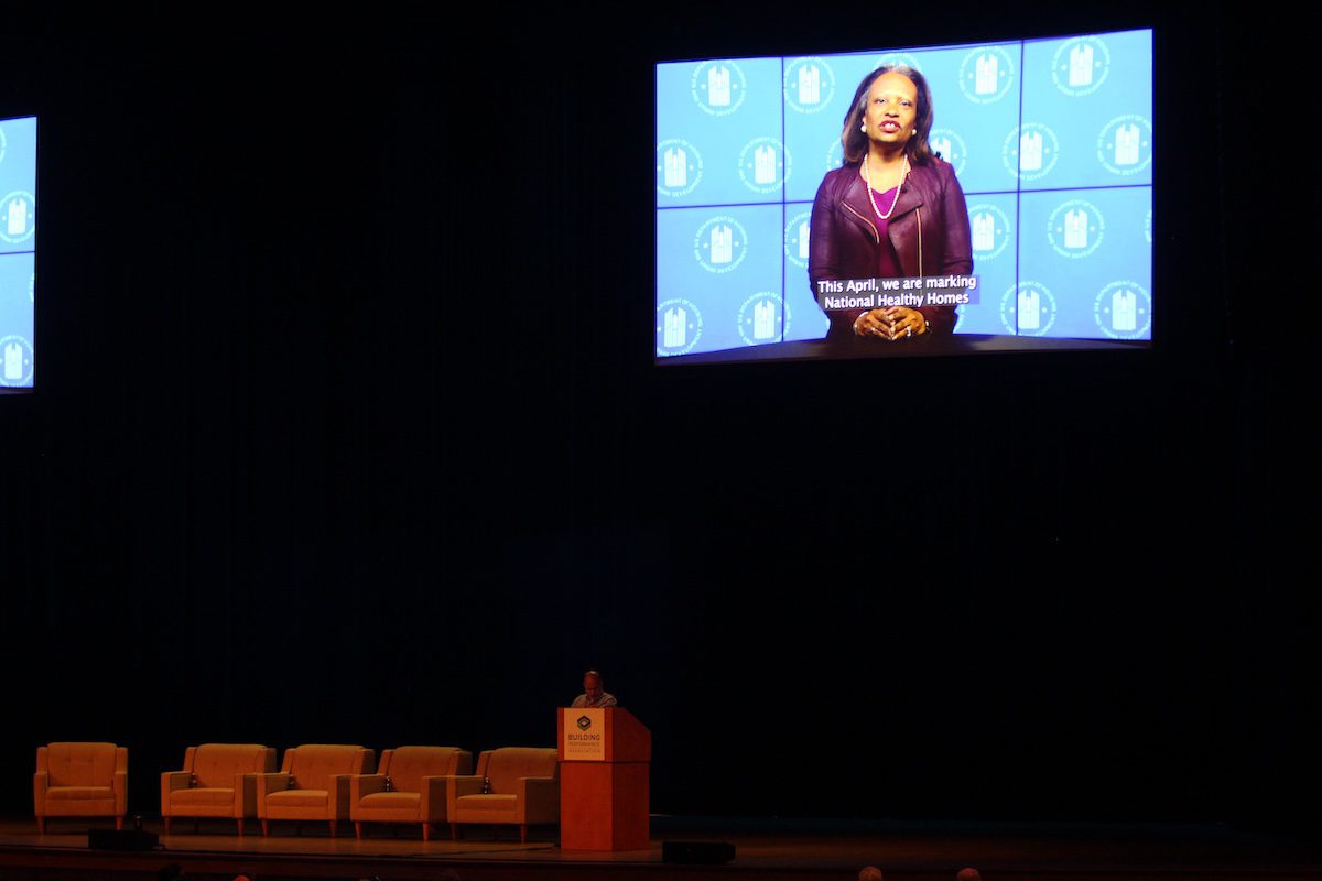 Image of a man presenting while a video of a woman plays in the background