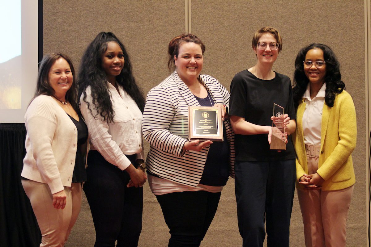 Image of 5 diverse women posing with a plaque and trophy