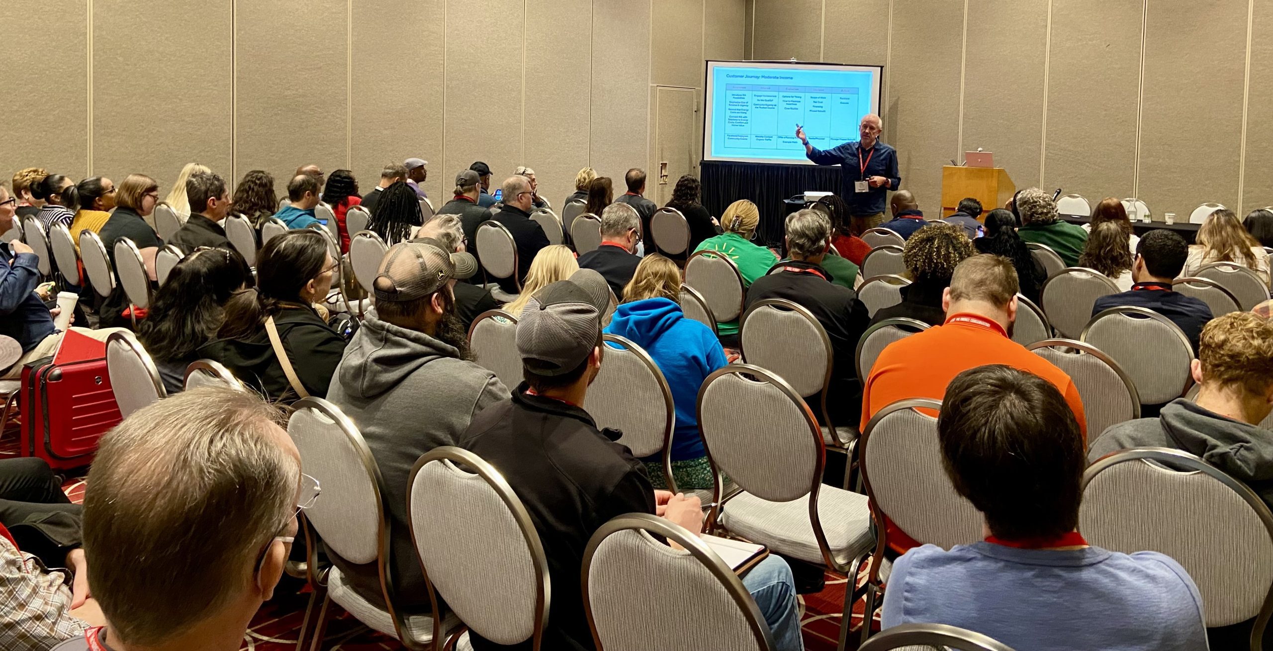 Image of a presentation being presented in a hotel with people listening and the presenter presenting.