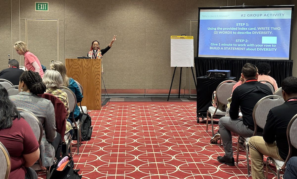 Image of a woman holding up a peace sign in front of a audience while giving a presentation