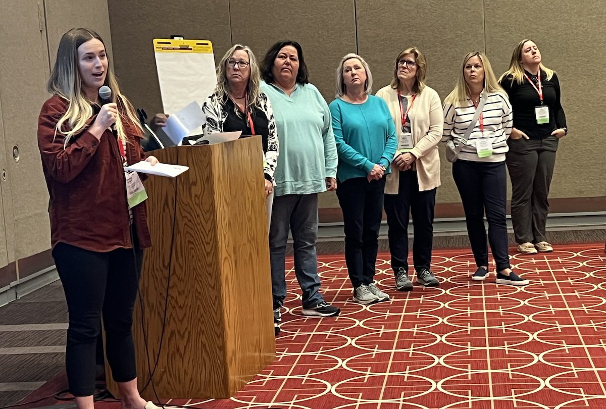 Image of a women presenting while 6 women stand beside her listening