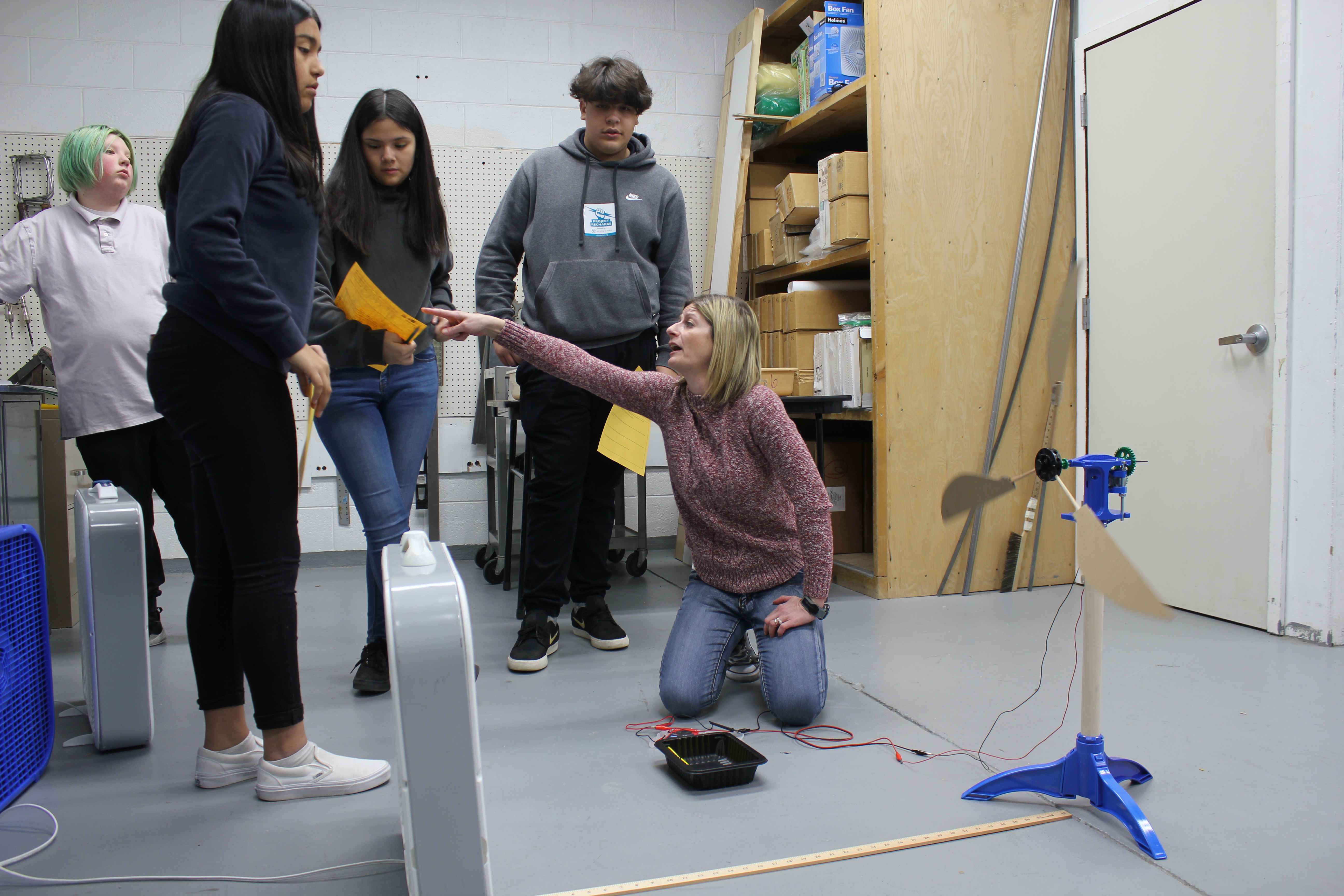 A woman kneels on the floor and points to a student to instruct them in a hands on activity. There are students surrounding the women kneeling. They are speaking to one another.