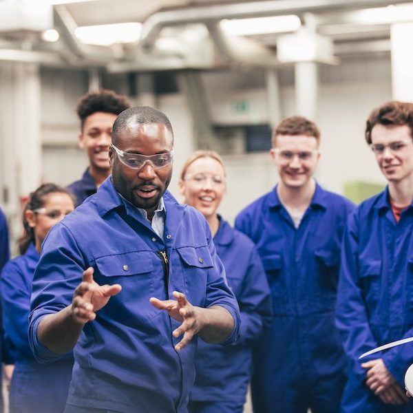 A tutor teaches his class about renewable energy in an engineering workshop. They are all wearing protective eyewear and blue coveralls.